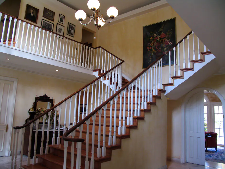 Interior view of a grand foyer with a staircase, white balusters, wood railings, and a chandelier. A painting hangs on the wall.
