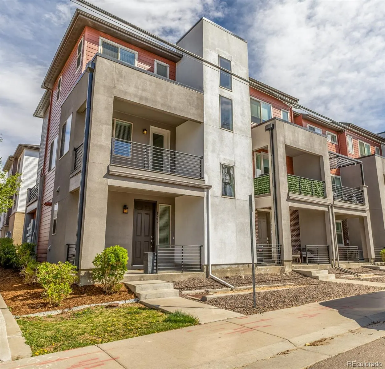 A modern townhouse with gray stucco and red siding, featuring balconies with metal railings and a small front yard.