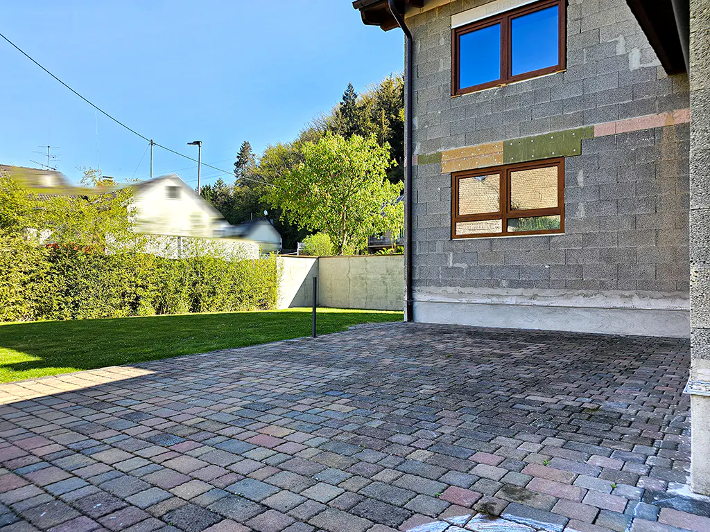 Exterior view of a gray block house with brown window frames and a brick patio. Green grass and trees are in the background.