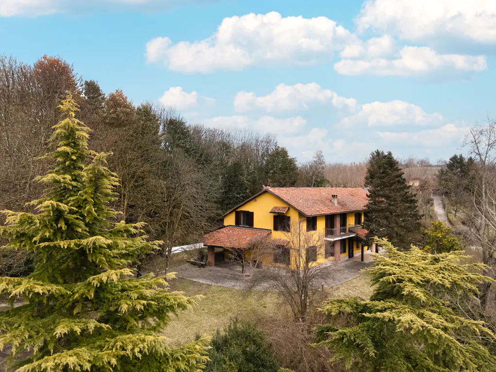 Two-story yellow house with a red tile roof surrounded by trees under a blue sky with white clouds.