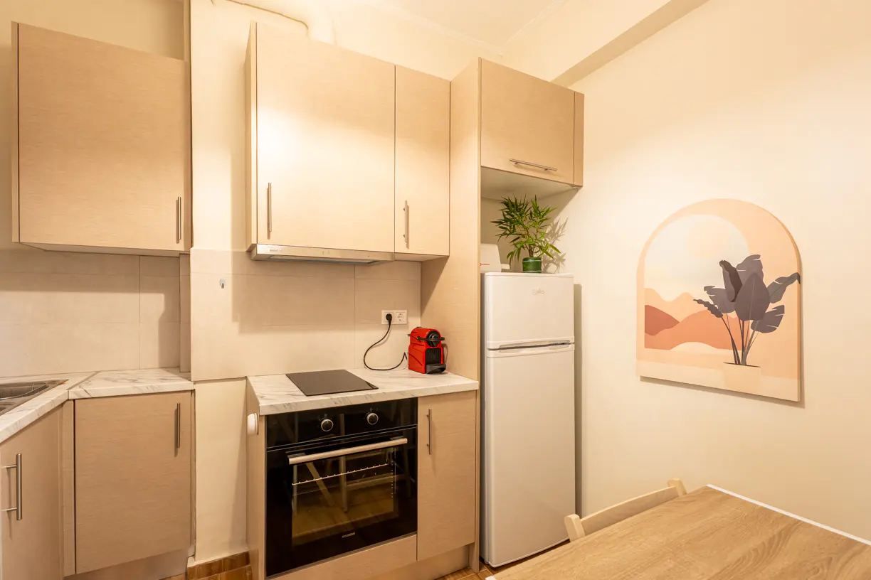 A compact kitchen with light wood cabinets, a white fridge, and a black oven. A red coffee maker sits on the counter. A plant is on top of the fridge. A desert landscape art piece hangs on the wall.