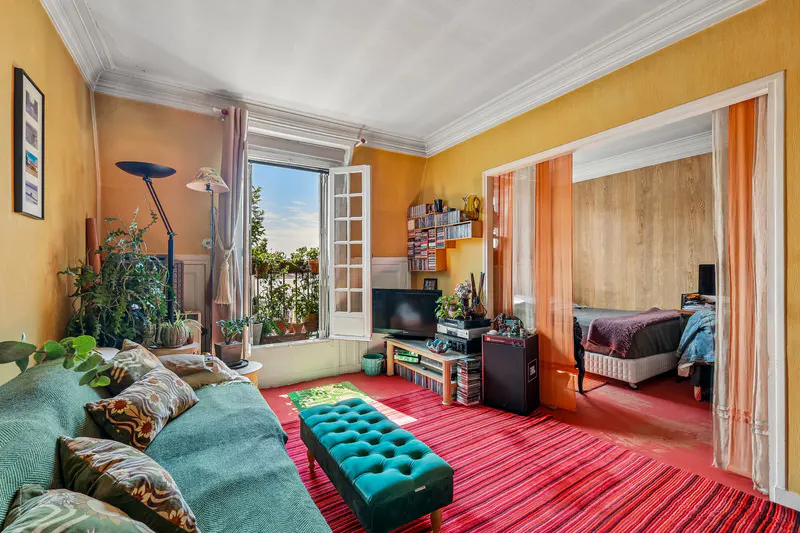 A cozy living room with yellow walls, a green sofa, and a red striped rug. An open balcony door shows plants and a view. A bedroom is visible through a doorway.