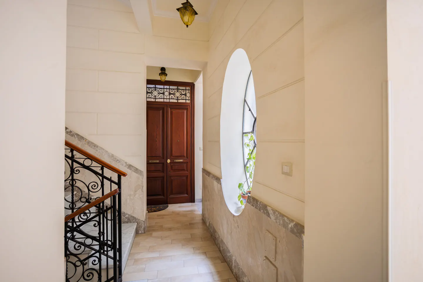 Hallway with cream walls, marble baseboards, a wrought iron staircase, and a unique oval window with a small plant.