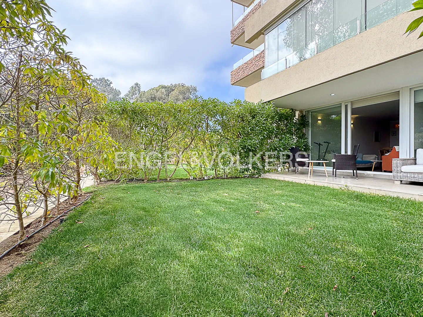 A green lawn leads to a modern apartment patio with outdoor furniture, framed by lush hedges and a multi-story building.