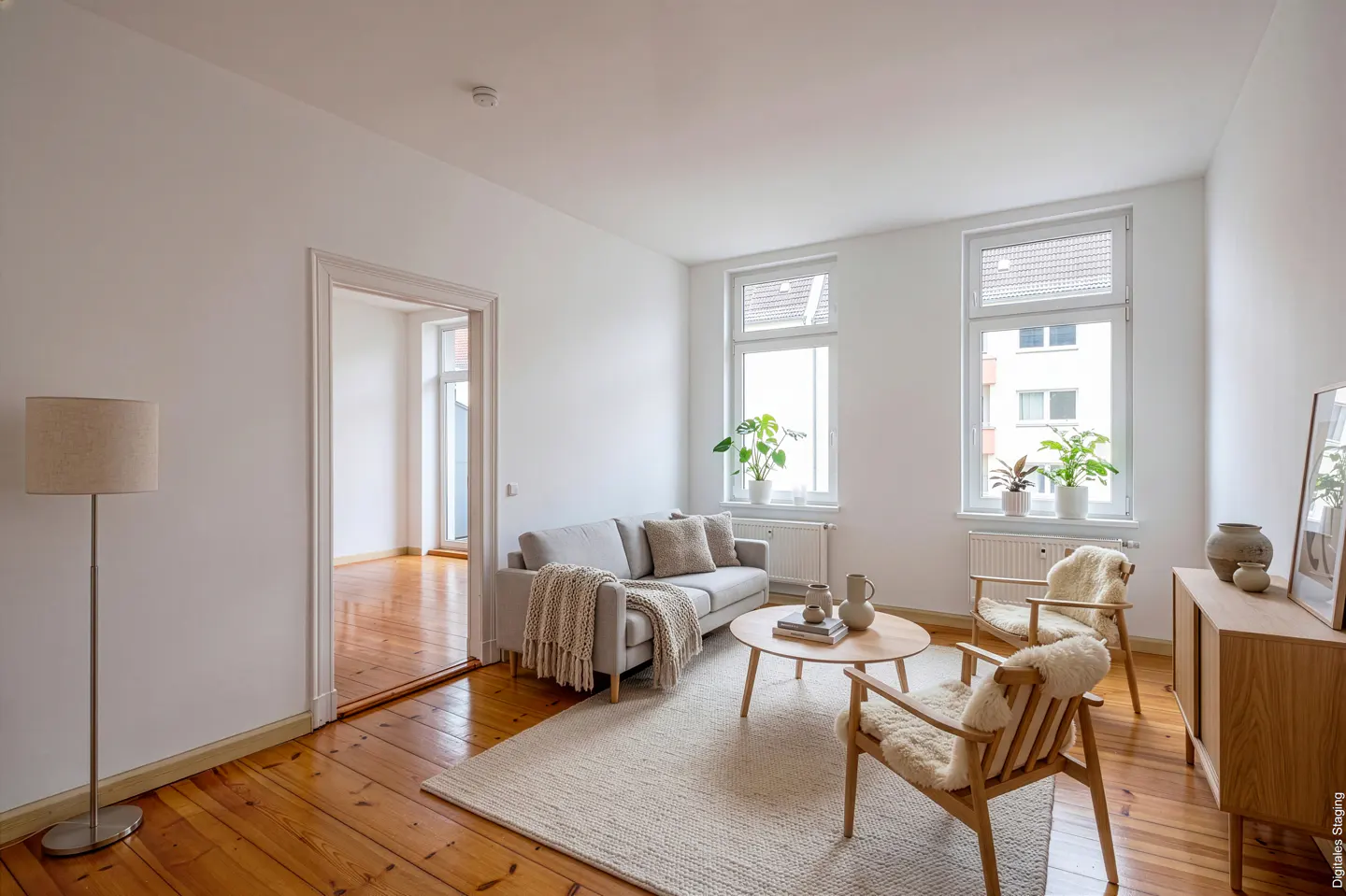 Bright living room with wood floors, white walls, and large windows. A gray sofa, round coffee table, and two chairs sit on a cream rug. Plants adorn the windowsills.