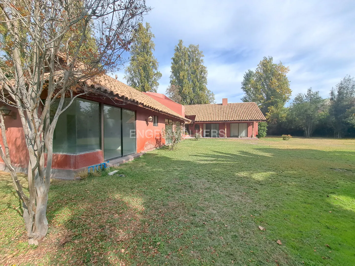 Exterior view of a red, single-story house with a brown tile roof and large windows, surrounded by a green lawn and trees.