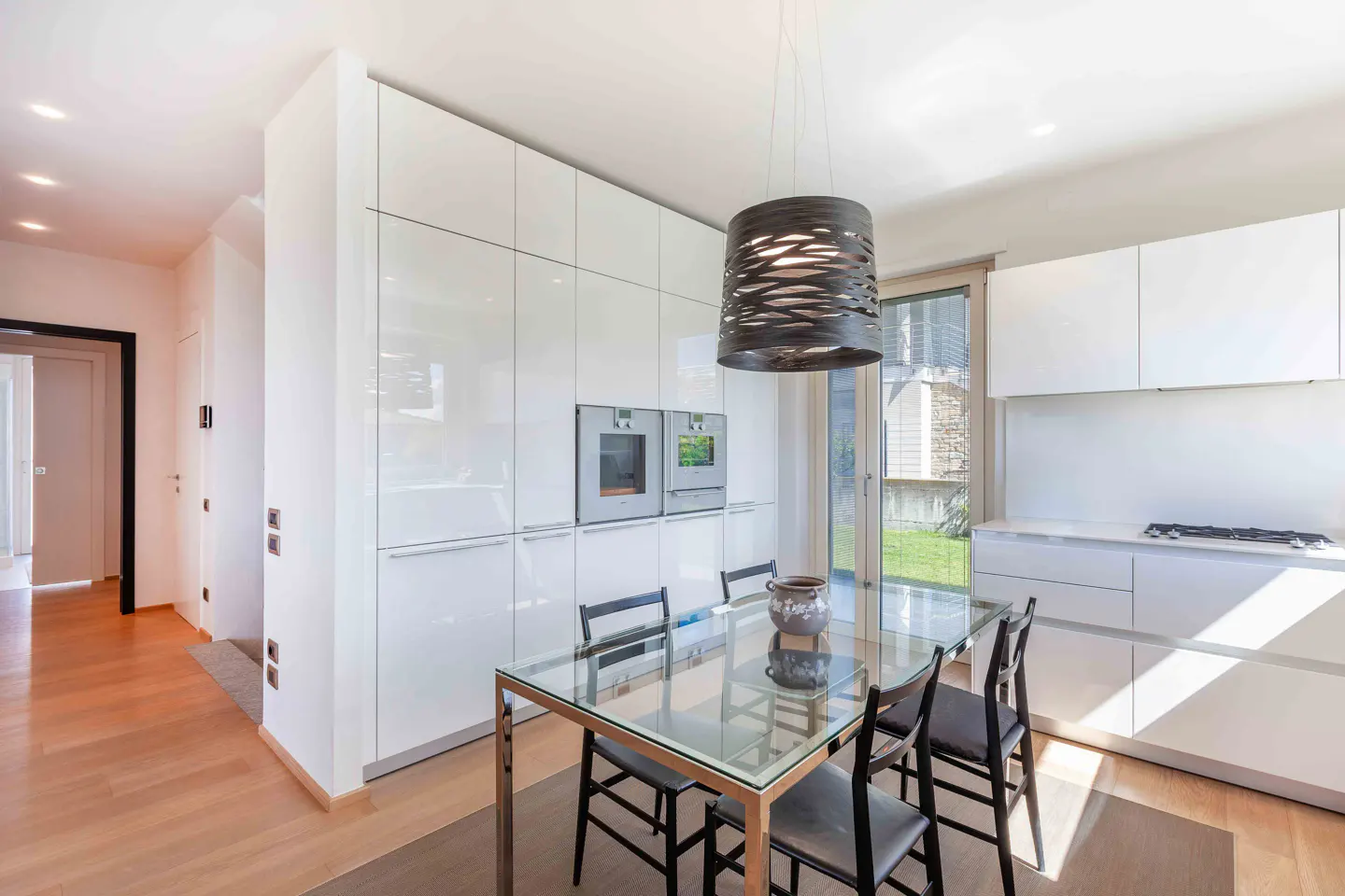 Bright kitchen with white cabinets, stainless steel appliances, and a glass dining table with black chairs. A modern light fixture hangs above the table.