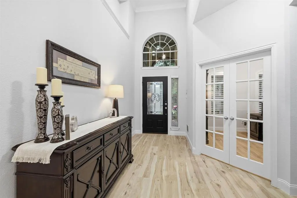 Bright foyer with light wood floors, white walls, and a black front door with a decorative window. A dark wood console table sits against the wall with candles and a lamp. White French doors are on the right.