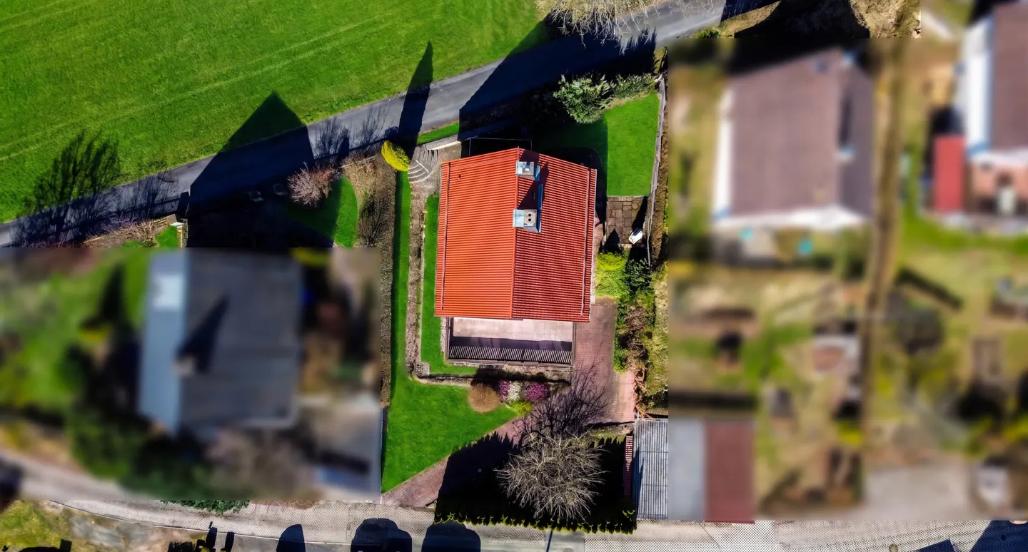 Aerial view of a house with a red roof, green lawn, and a paved patio. A road runs alongside the property.