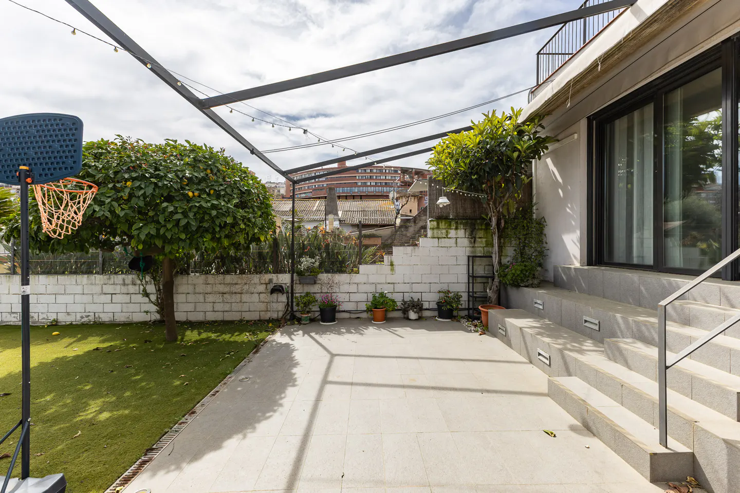 Backyard with a basketball hoop, grass, and a tiled patio. Steps lead to a modern house with a black-framed sliding glass door. Overhead metal pergola.