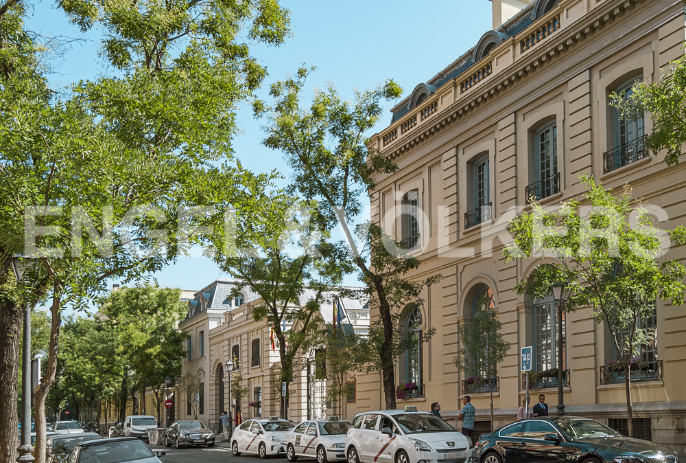 Street view of beige buildings with blue window frames, green trees, and parked cars on a sunny day.