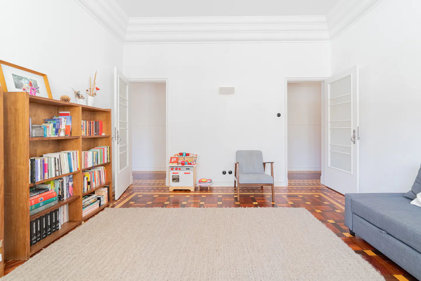 Bright living room with white walls, parquet floor, and beige rug. A bookcase, gray chair, and toy kitchen are visible. Two open doorways lead to other rooms.
