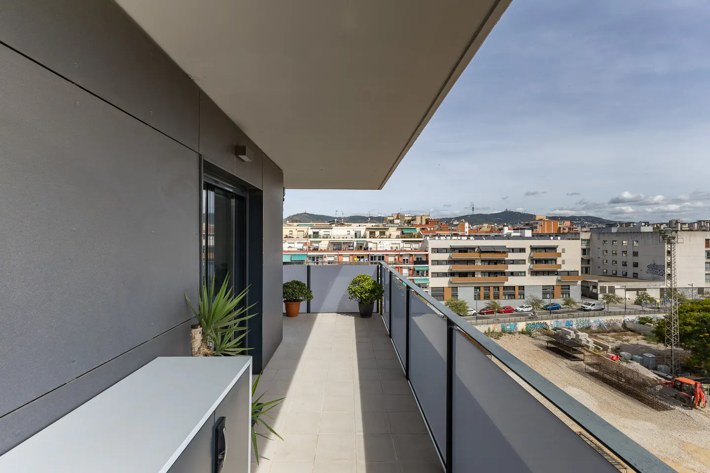 Modern apartment balcony with gray walls, potted plants, and a glass railing overlooking a city street and distant hills.