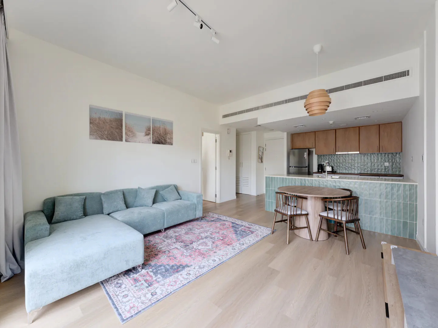 Bright, open-concept apartment with a blue sectional sofa, patterned rug, and a kitchen with wood cabinets and a round dining table.