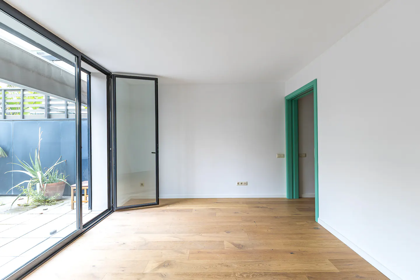 Empty room with light wood floors, white walls, a green door frame, and large glass doors opening to a patio with plants.