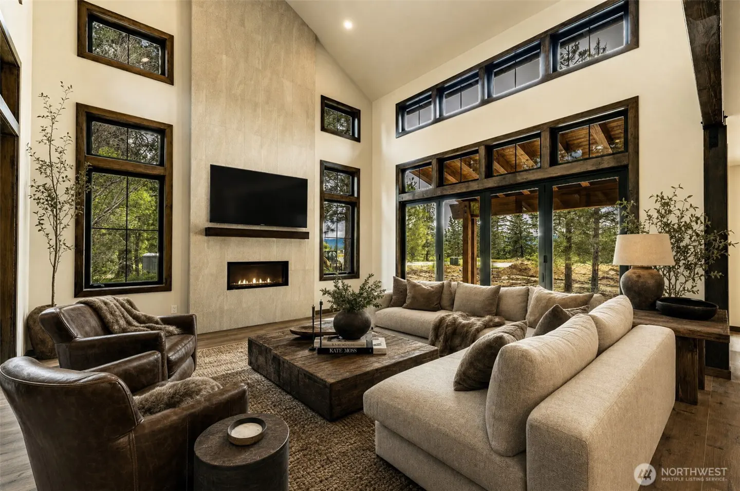 Living room with high ceilings, a fireplace, and large windows. Two brown leather chairs face a large, square, wooden coffee table and a beige sectional sofa.
