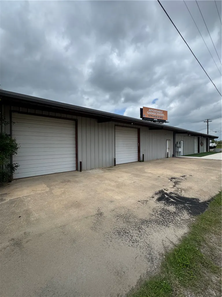 A long, grey metal commercial building with two large white garage doors and a billboard under a cloudy sky.