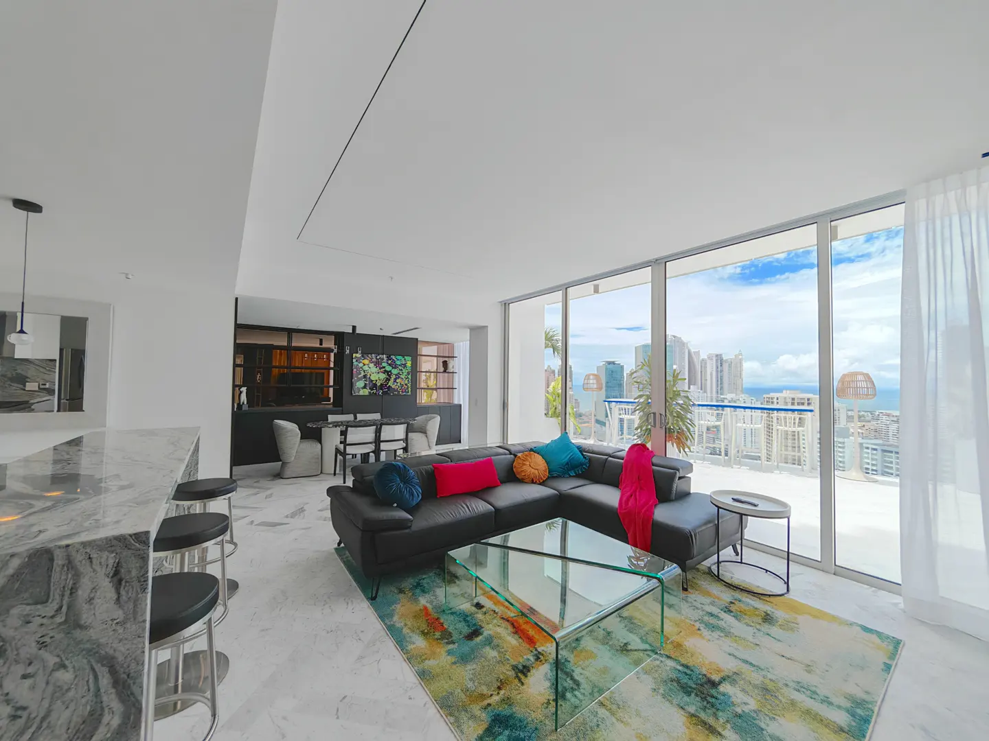 Bright, modern living room with a black leather sectional sofa, colorful pillows, and a glass coffee table. Large windows offer a city view.