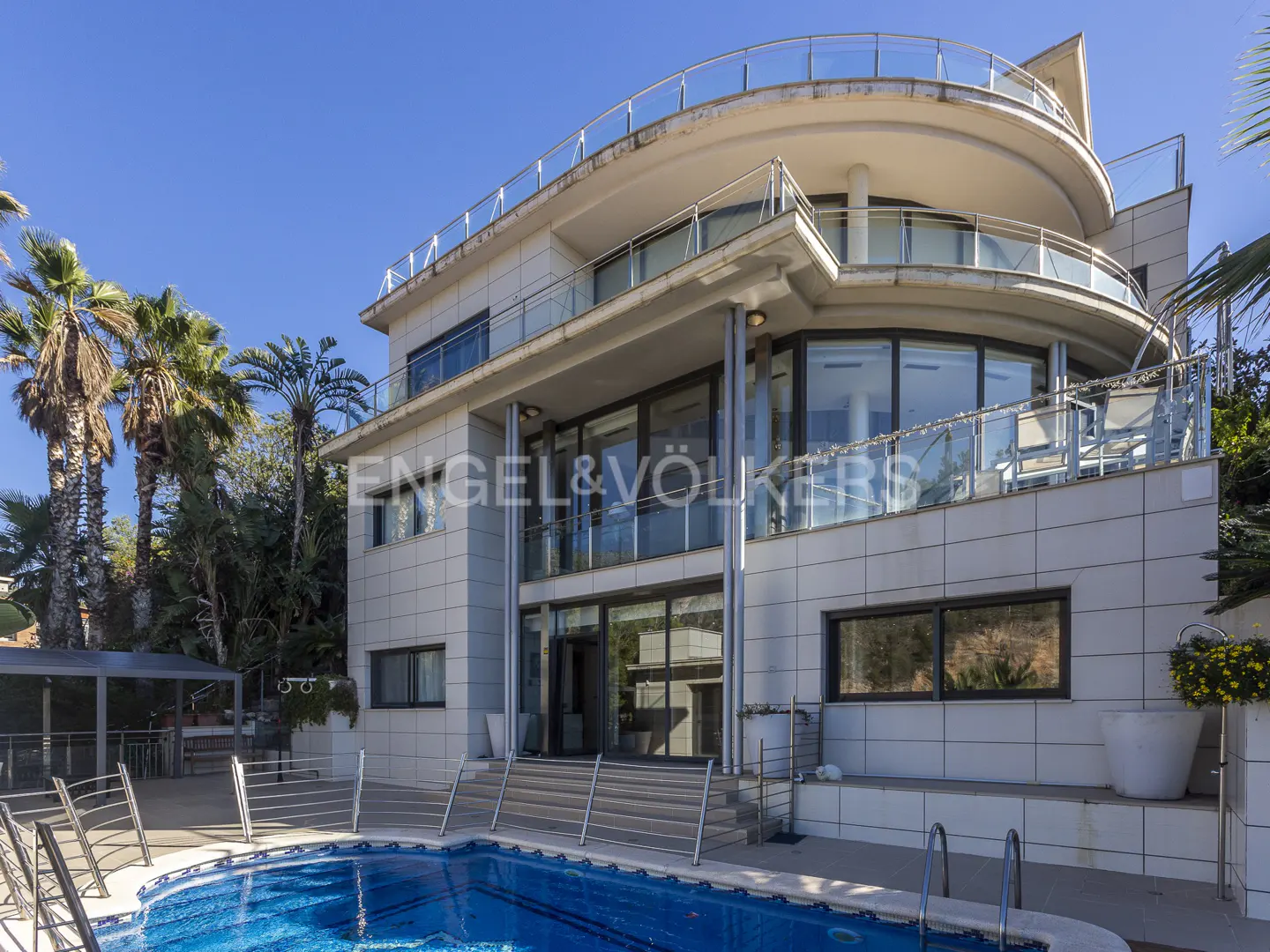 Exterior view of a modern, multi-story house with a blue tiled pool in the foreground and palm trees in the background.
