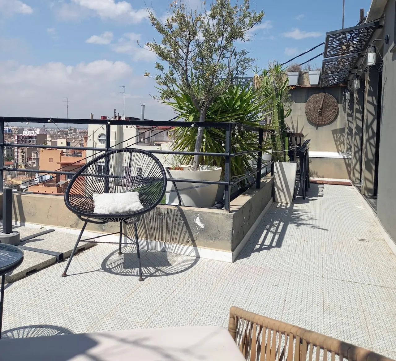 Rooftop patio with black chair, white pillow, potted plants, and city views. The floor is white tile and the sky is blue.