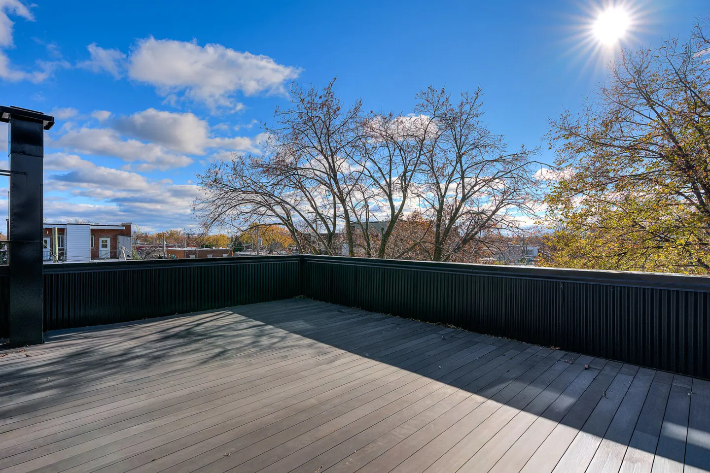 Outdoor deck with gray wood planks and black railing, under a blue sky with white clouds and a bright sun. Trees in the background.