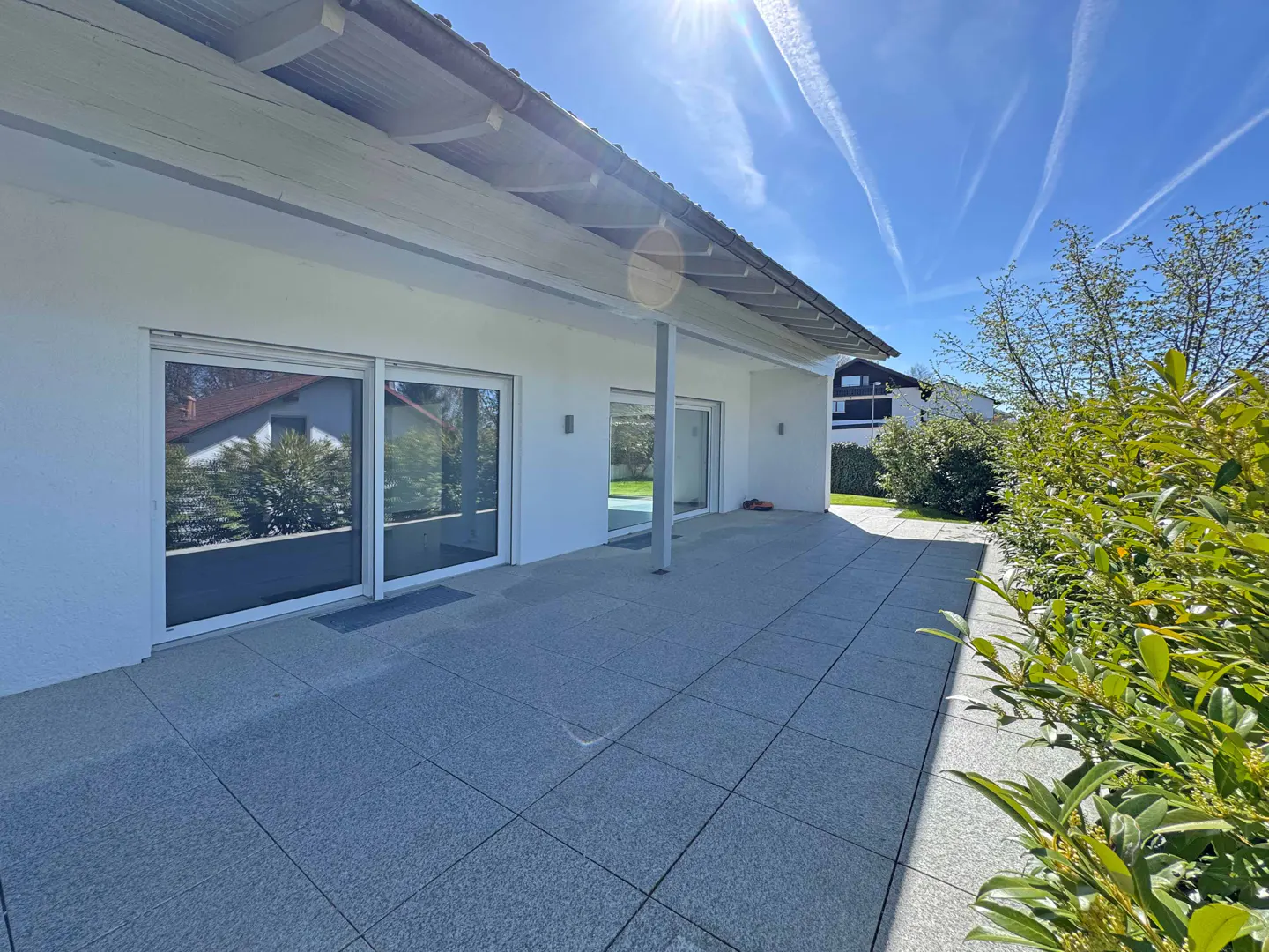 Exterior view of a white house with a gray tiled patio, sliding glass doors, and green bushes under a sunny blue sky.