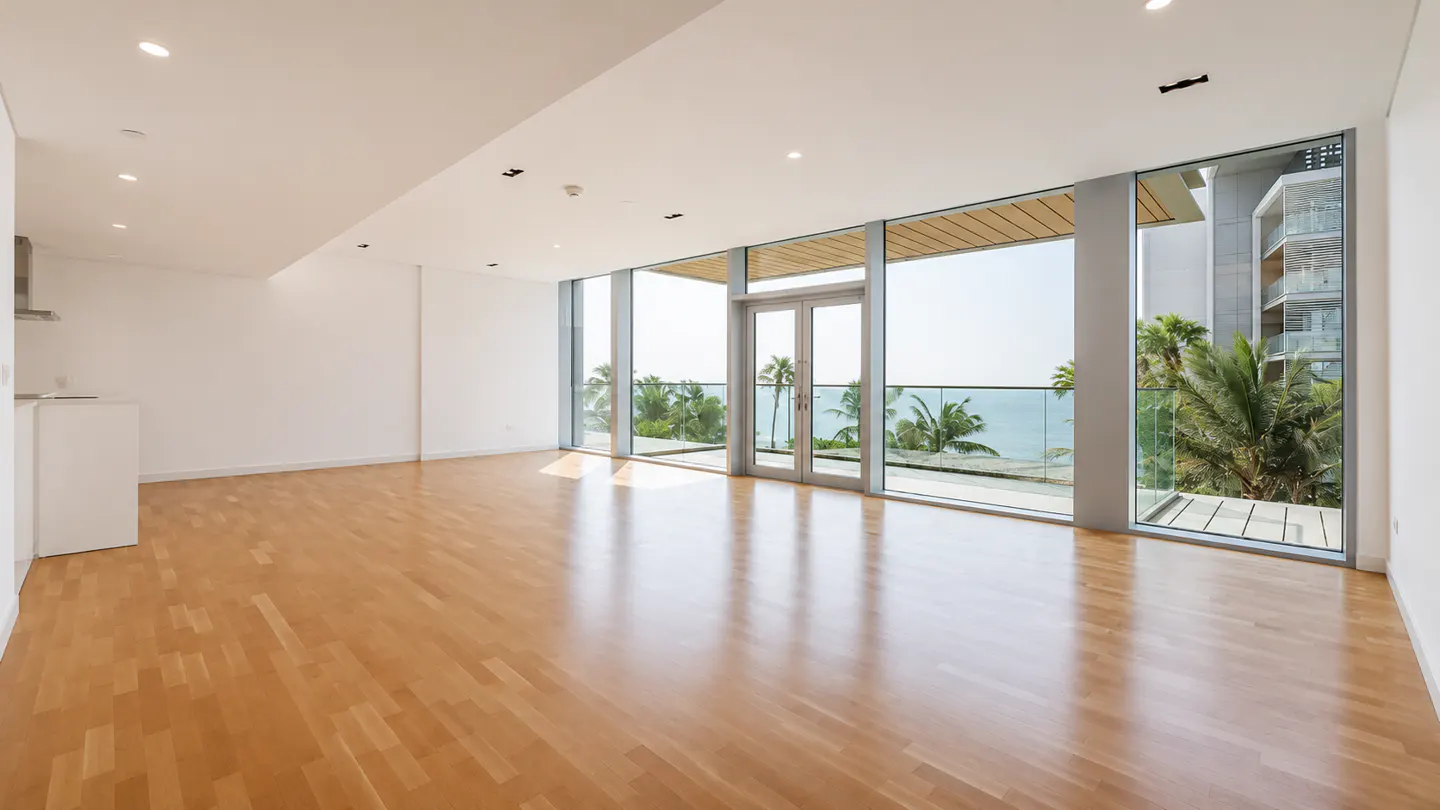 Bright, empty room with light wood floors and white walls. Large windows offer ocean views with palm trees. Double doors lead to a balcony.