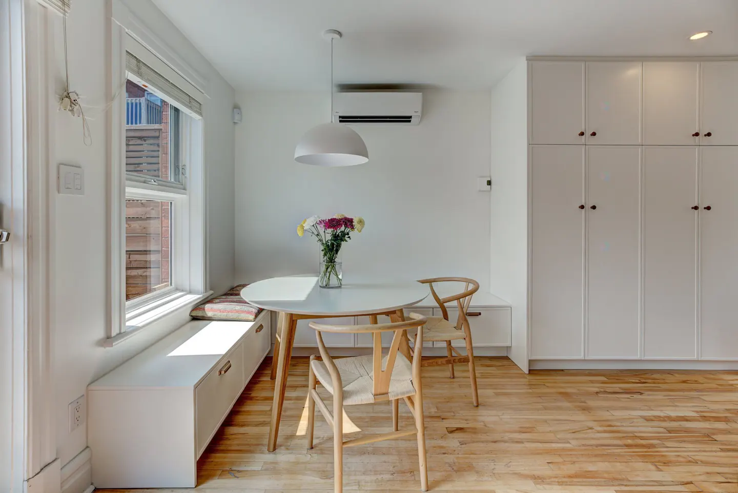 Bright dining area with a round white table, two wooden chairs, and a vase of flowers. A window seat and white cabinets add storage.