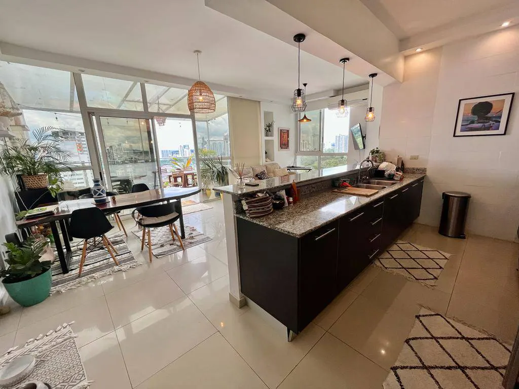 A bright, modern apartment interior with a kitchen island, dining table, and floor-to-ceiling windows overlooking a cityscape.