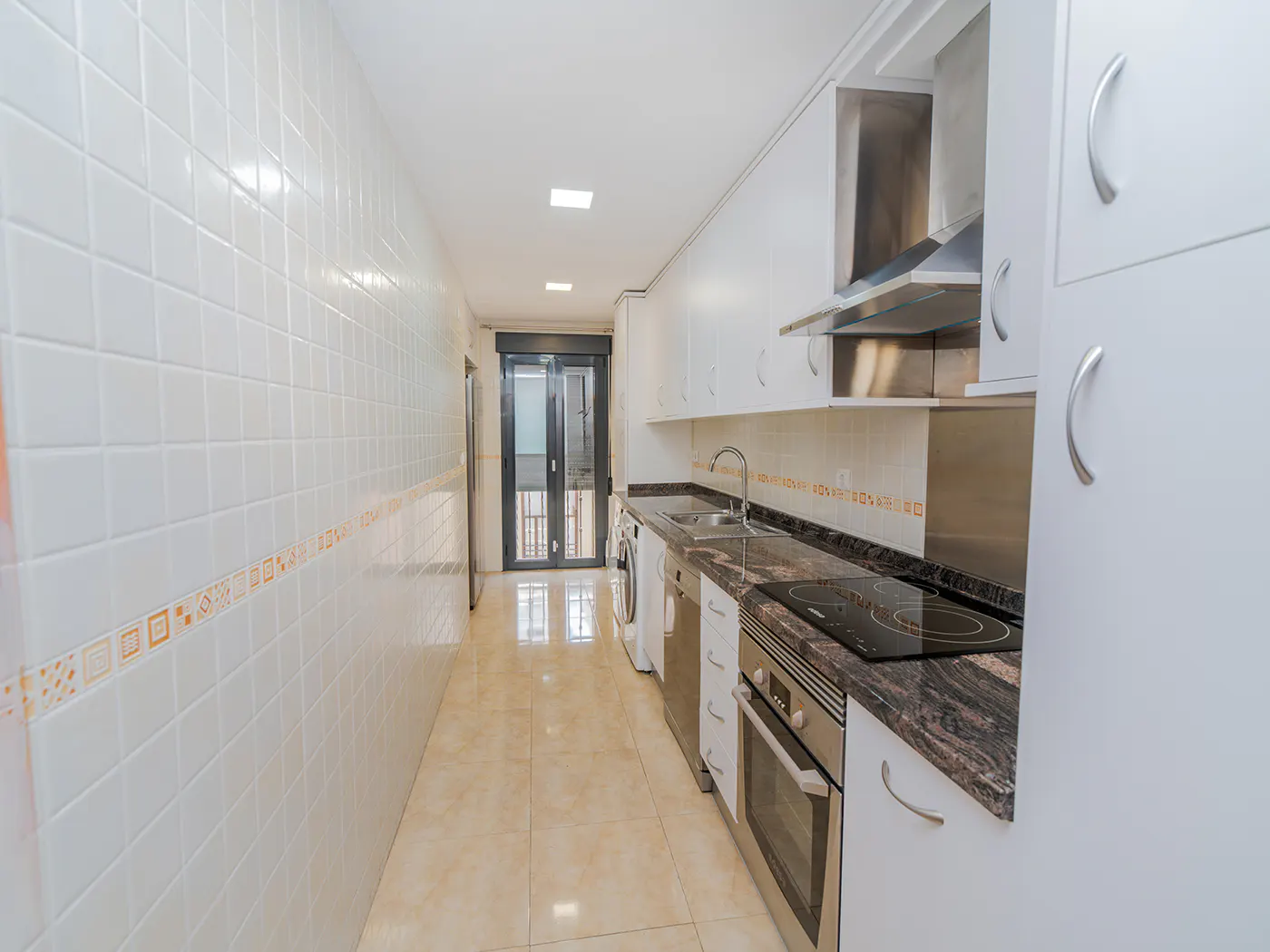 A long, narrow kitchen with white cabinets, stainless steel appliances, and a tiled floor. A door leads to a balcony.