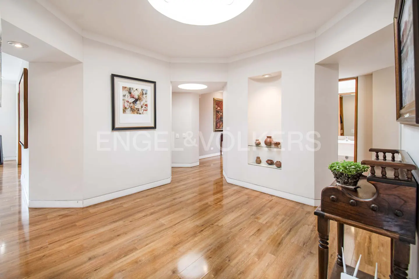 Hallway with light wood floors, white walls, recessed lighting, and artwork. A dark wood table with a plant sits in the foreground.