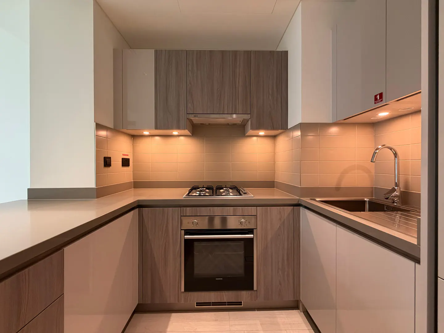 Modern U-shaped kitchen with wood-grain cabinets, stainless steel oven, gas stovetop, and warm under-cabinet lighting.