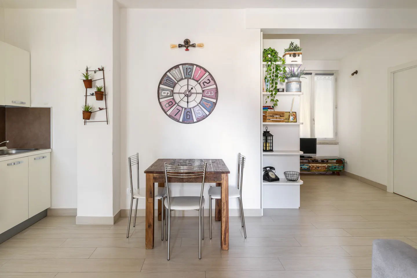 Bright apartment interior with a wooden dining table, four chairs, and a colorful clock on a white wall. A kitchen and living room are visible.