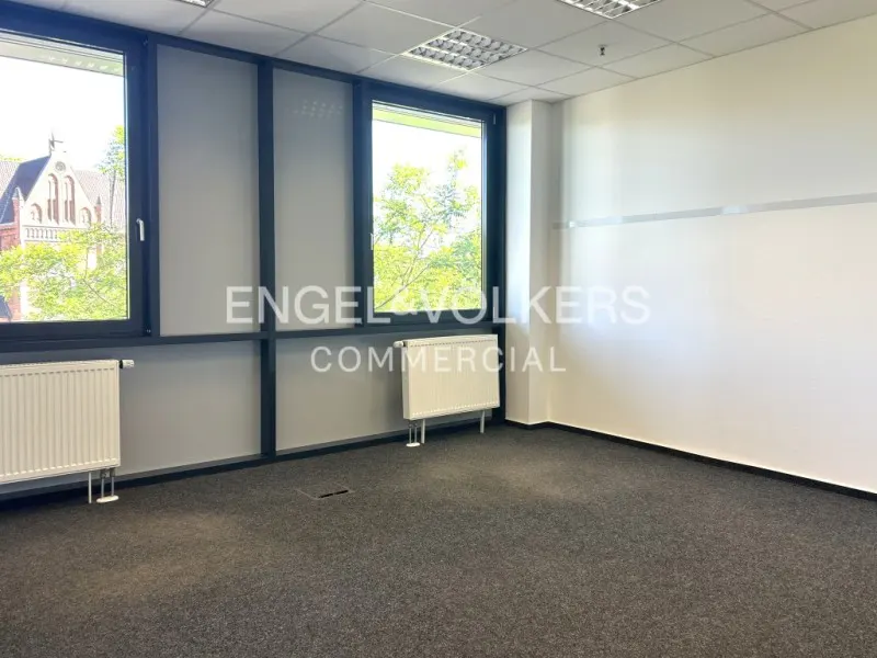 Empty office space with gray carpet, white walls, and black window frames. Radiators are under the windows, and a building is visible outside.