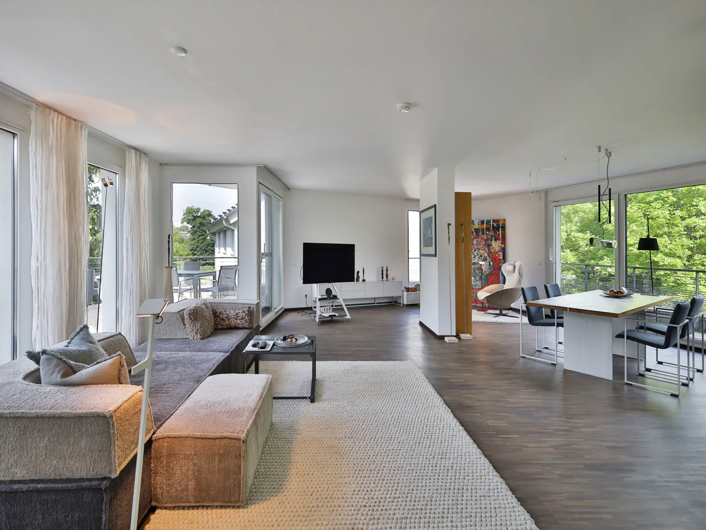 Open-concept living and dining area with a gray sectional sofa, white rug, and dining table with black chairs. Large windows offer views of greenery.