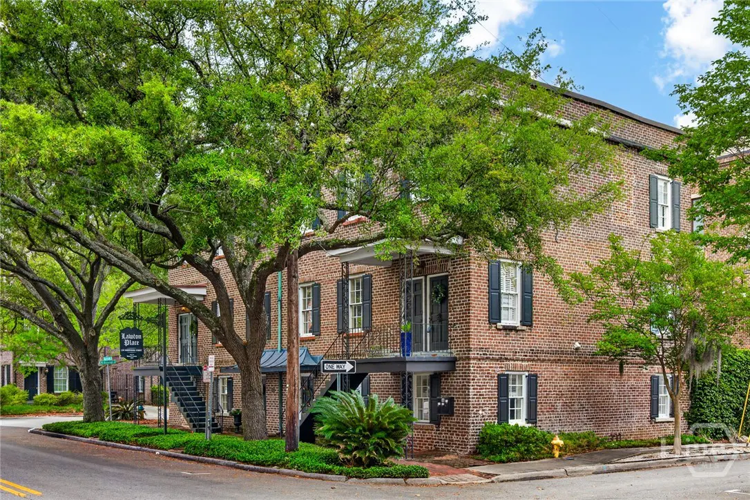 Exterior view of a two-story brick building with black shutters and wrought iron balconies, partially shaded by large trees.