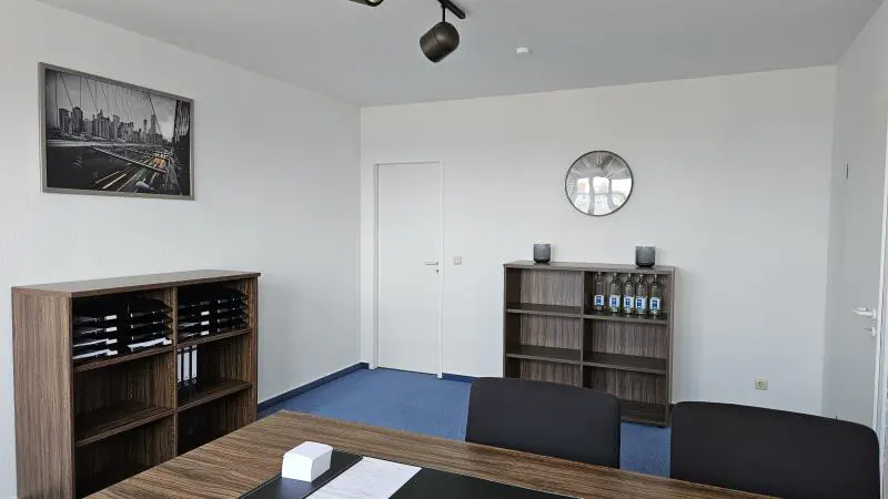 Office interior with blue carpet, white walls, and wood furniture. A framed city print hangs above a shelf with files. A clock and water bottles sit on another shelf.