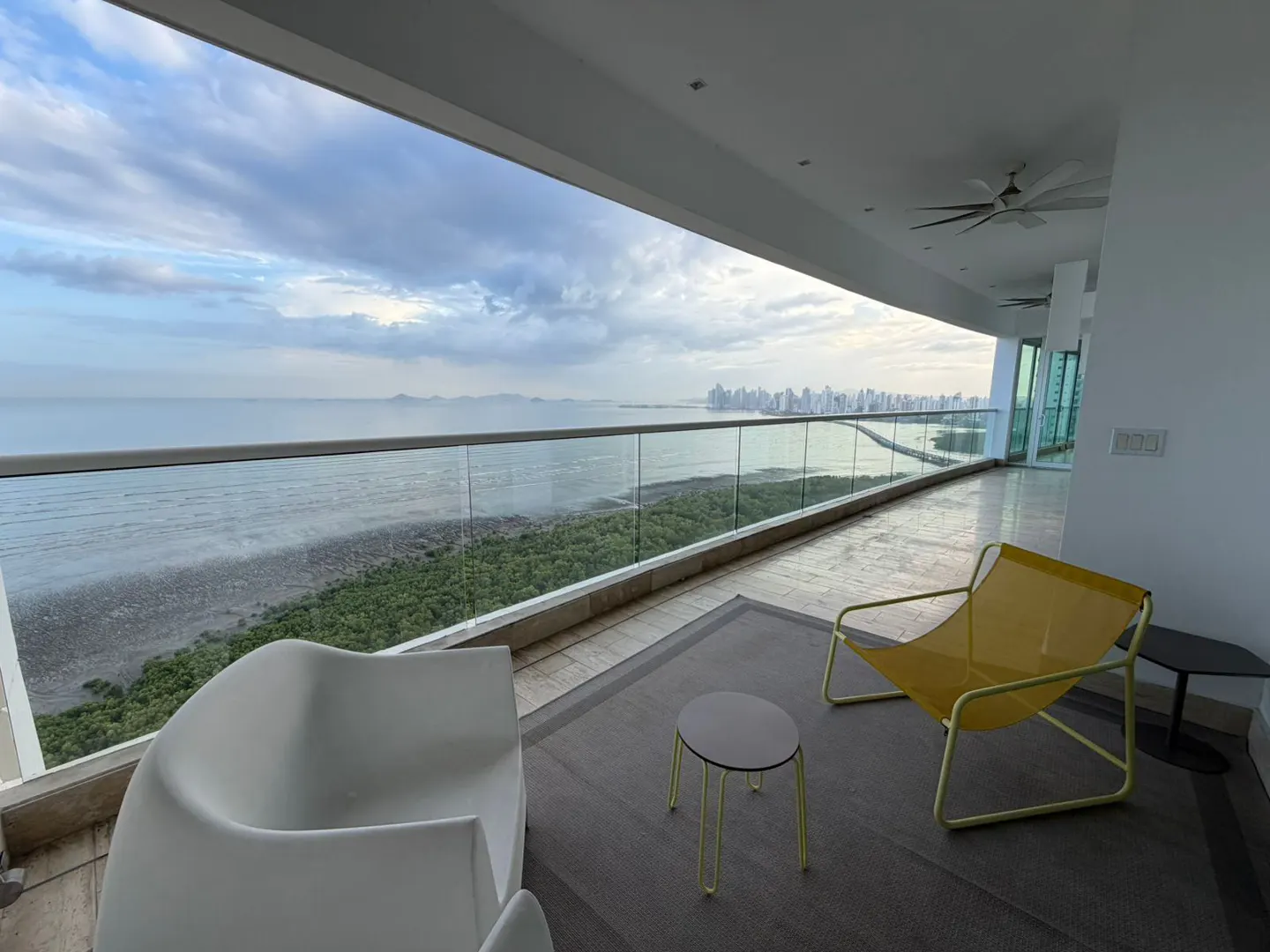 Balcony view with white chairs, yellow lounge, and city skyline. Ocean and green trees visible beyond glass railing.