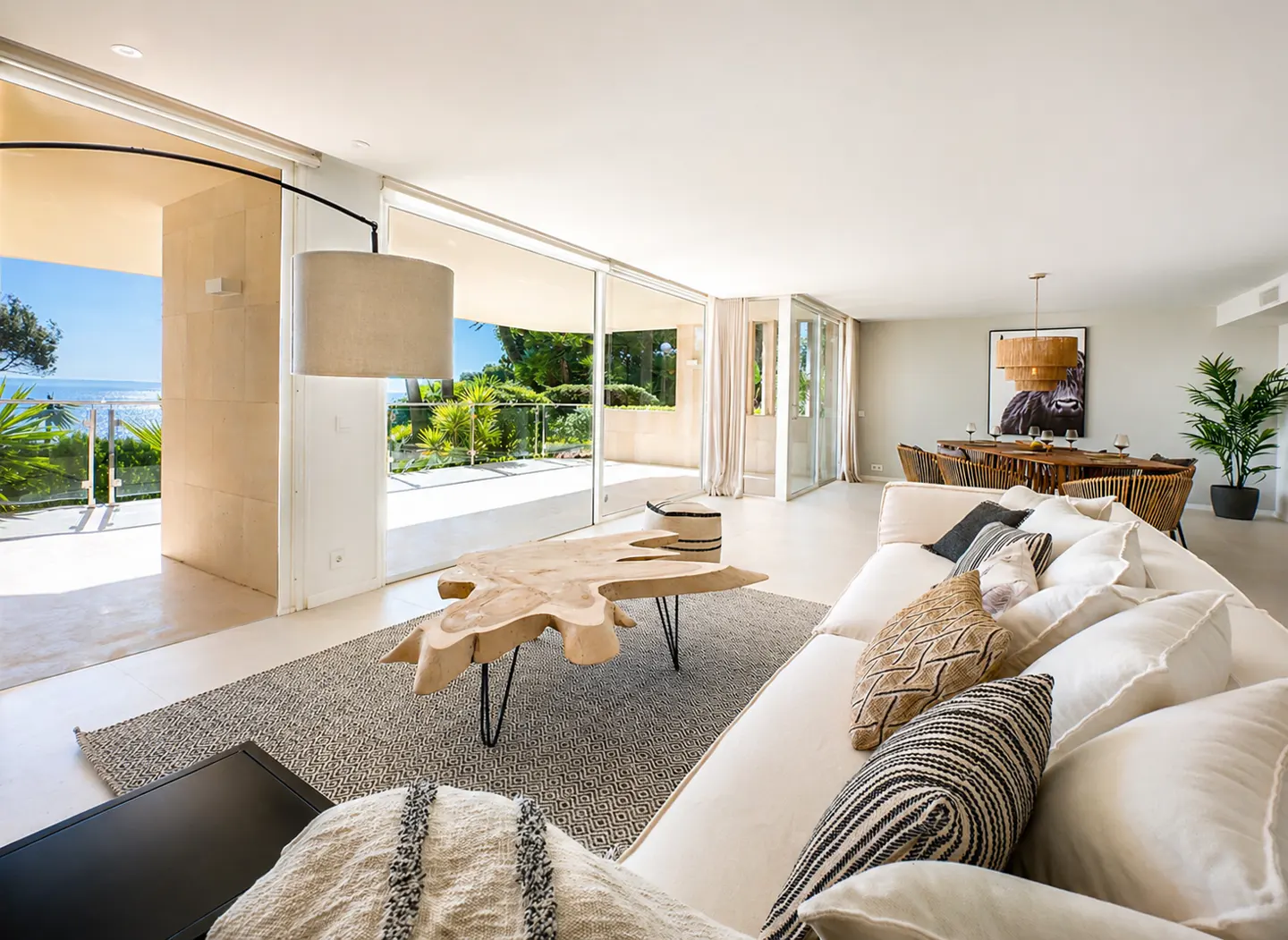 Bright living room with a white sofa, patterned pillows, and a unique wood coffee table. Sliding glass doors open to a patio with ocean views.