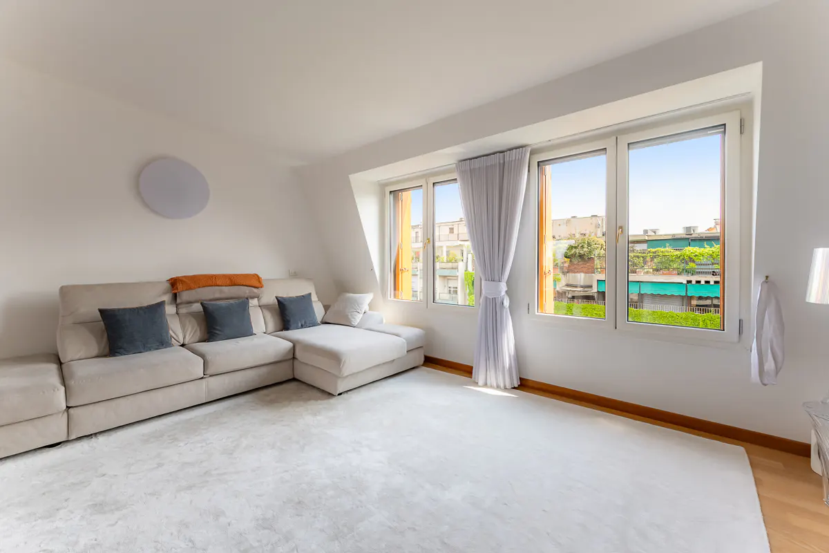 Bright living room with a beige sectional sofa, blue pillows, and a large window with a view of the city.