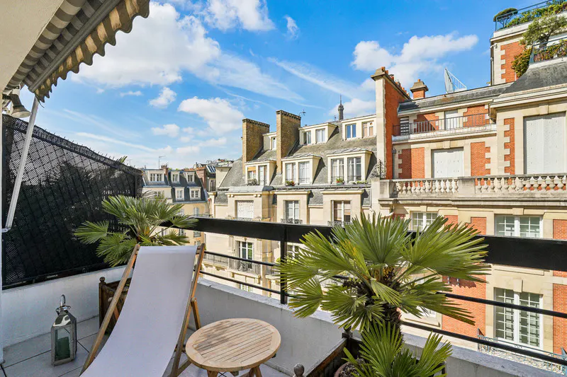 Balcony view with a white lounge chair, round table, and potted palm trees. Parisian buildings are visible in the background under a blue sky.