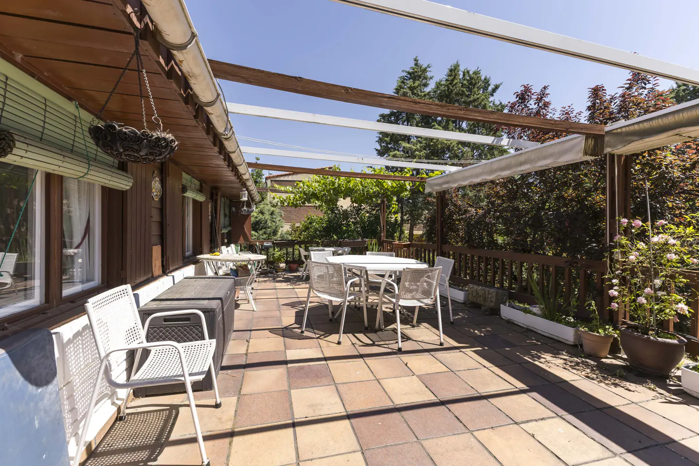 Outdoor patio with white metal chairs and tables on a tiled floor. A wooden pergola covers the area, with greenery and trees in the background.