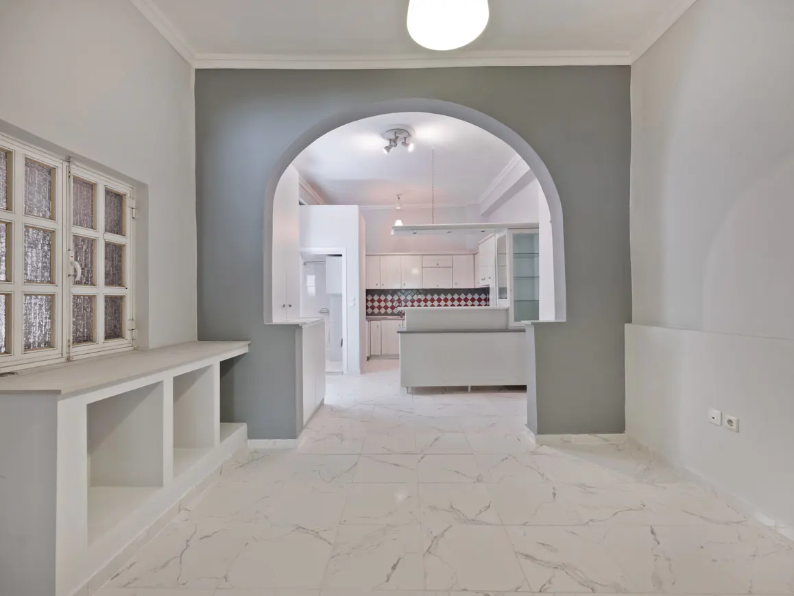 A bright, empty room with white marble floors and a gray archway leading to a white kitchen. A window with a shelf is on the left.