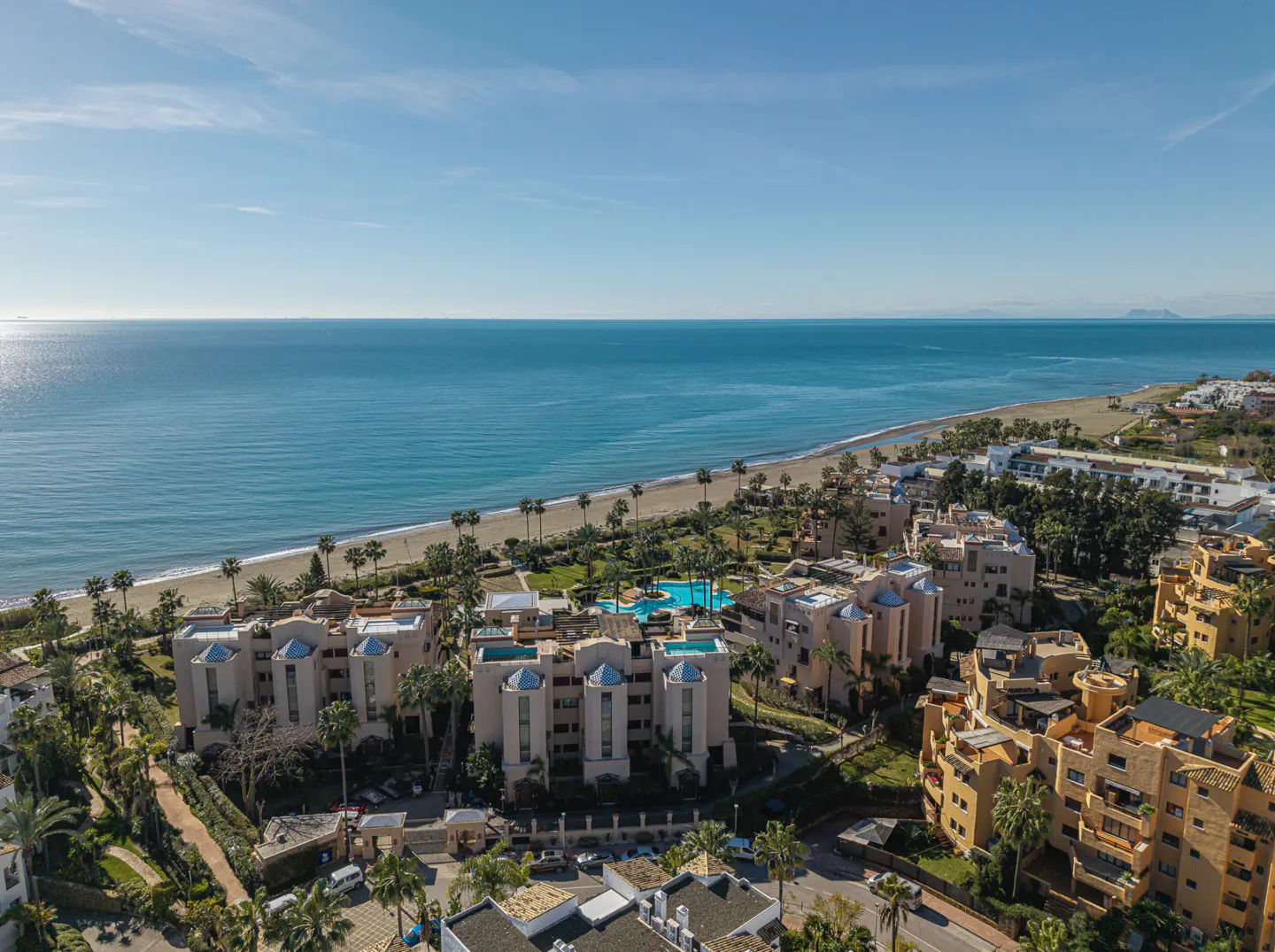 Aerial view of beachfront condos with palm trees, a sandy beach, and the blue ocean under a clear sky.
