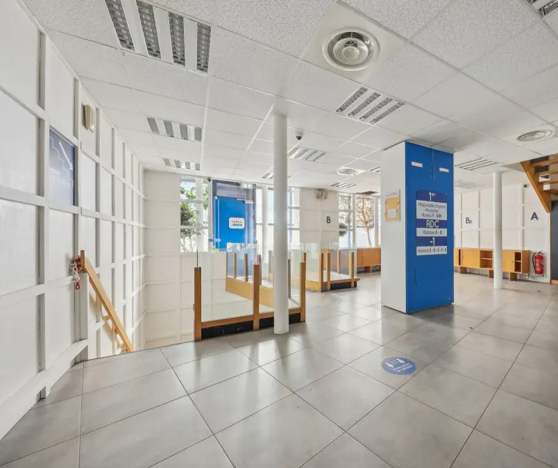 Bright office space with white walls, gray tile floor, and a blue directory sign. Stairs lead down on the left. Natural light from windows.