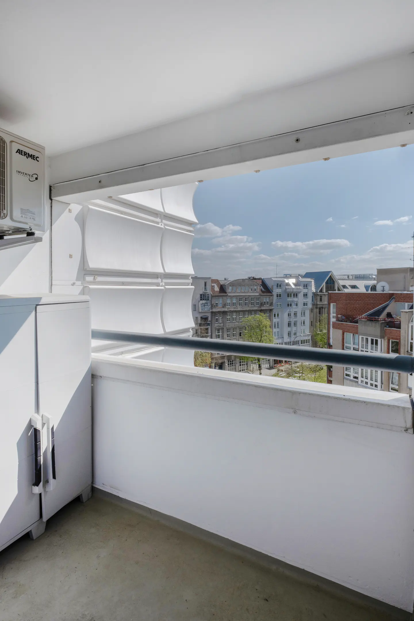 Balcony view with white walls, a metal railing, and a city skyline in the background under a blue sky.