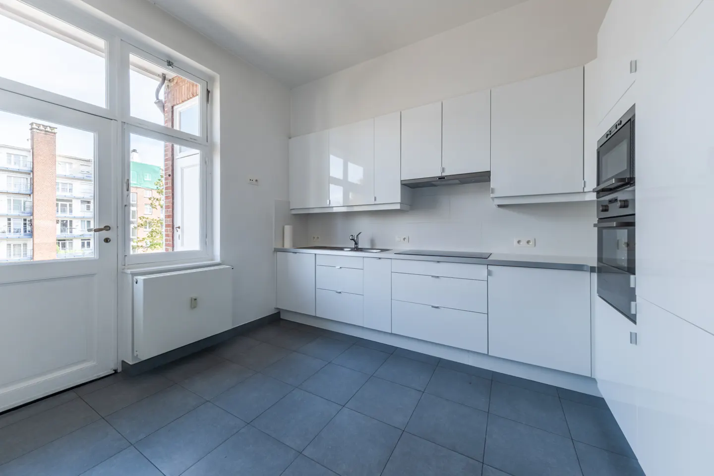 Bright kitchen with white cabinets, gray countertops, and gray tile floor. A white door leads to a balcony with a view of buildings.