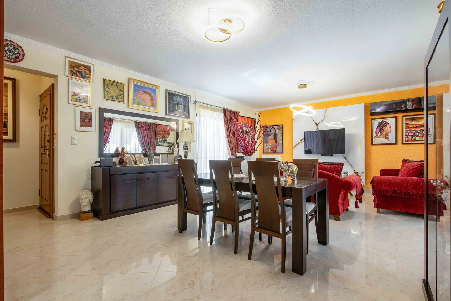 Dining area with dark wood table and chairs, marble floors, red sofa, and walls decorated with numerous framed artworks.