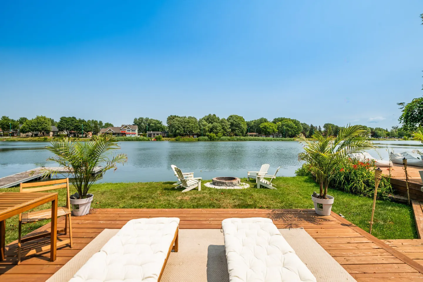 Waterfront property with a wooden deck, lounge chairs, fire pit, and lake view under a clear blue sky.