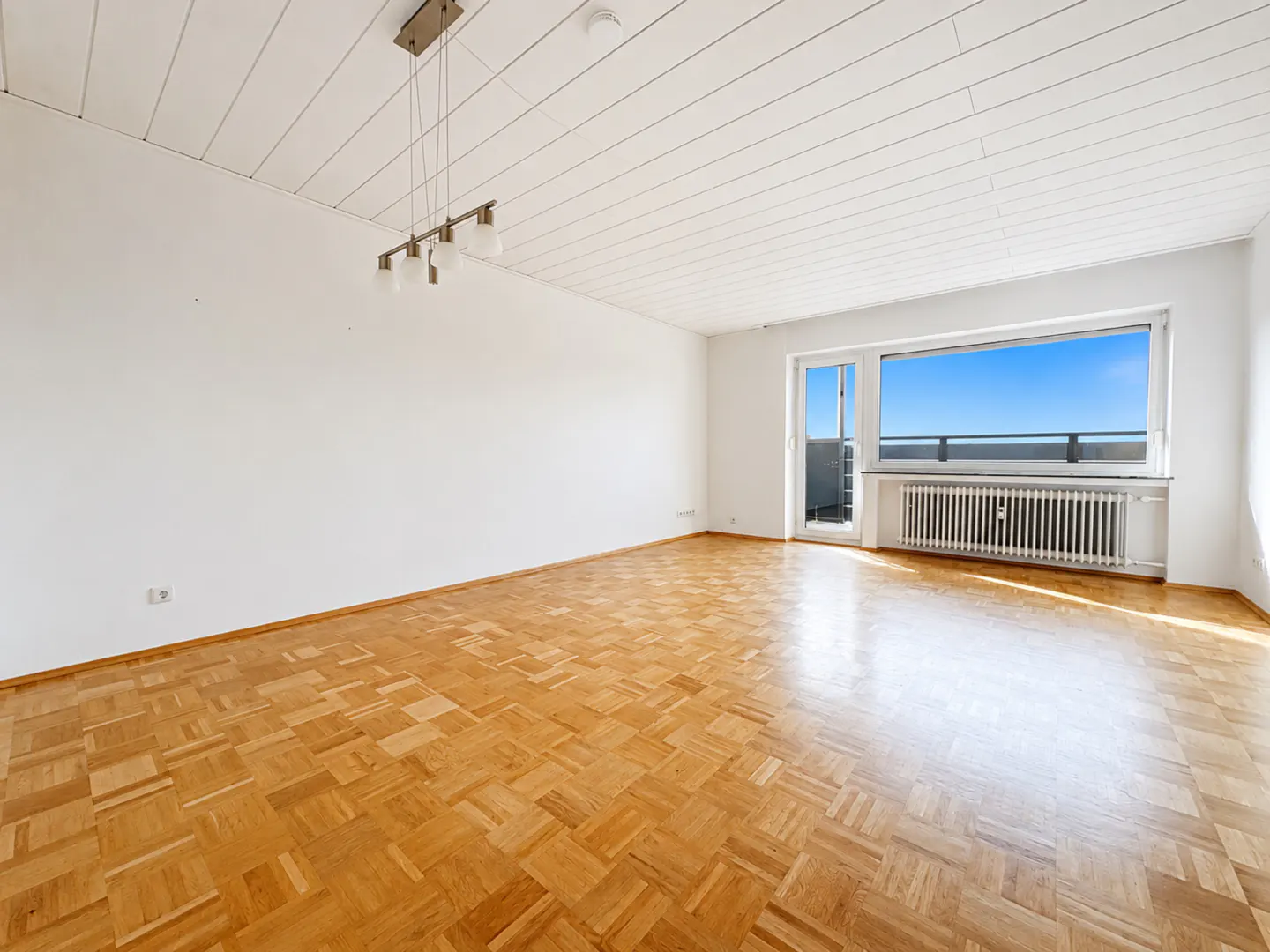Bright, empty room with parquet floors, white walls, and a large window with a balcony view of a clear blue sky.